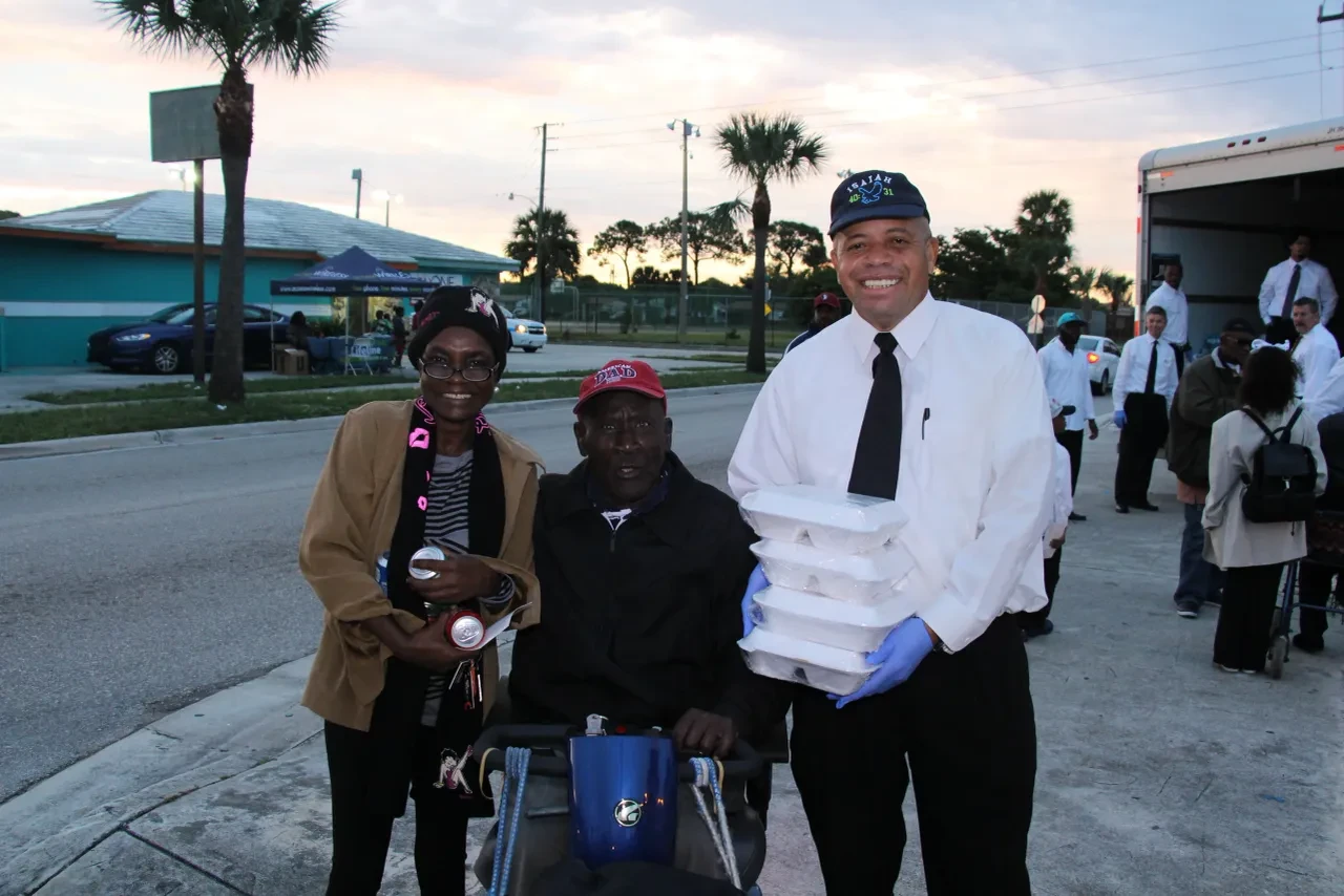 Volunteer handing food containers to community member in wheelchair at dusk