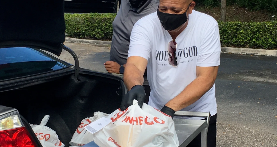 Masked volunteer in House of God shirt loading Golden Corral bags into car trunk