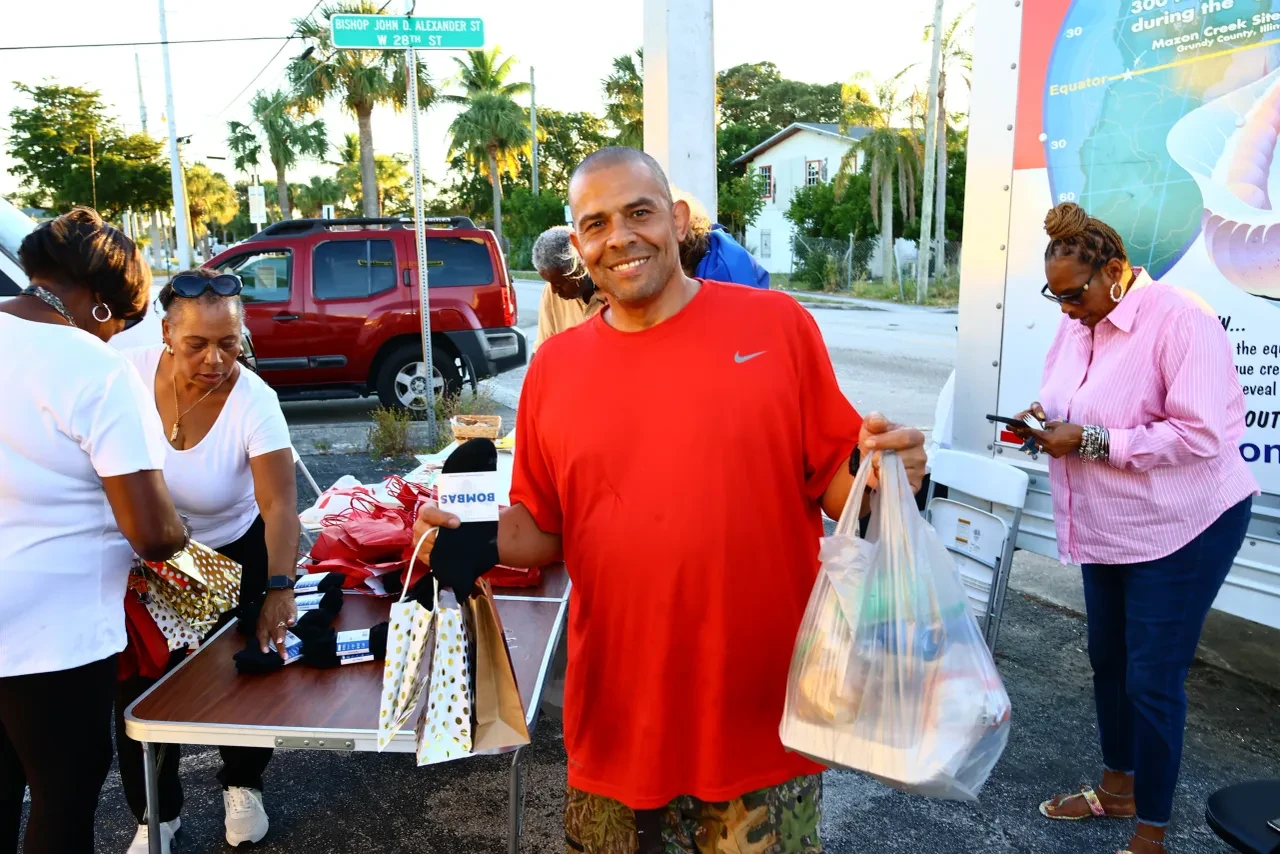 Man in red Nike shirt holding grocery bags at outdoor community distribution table