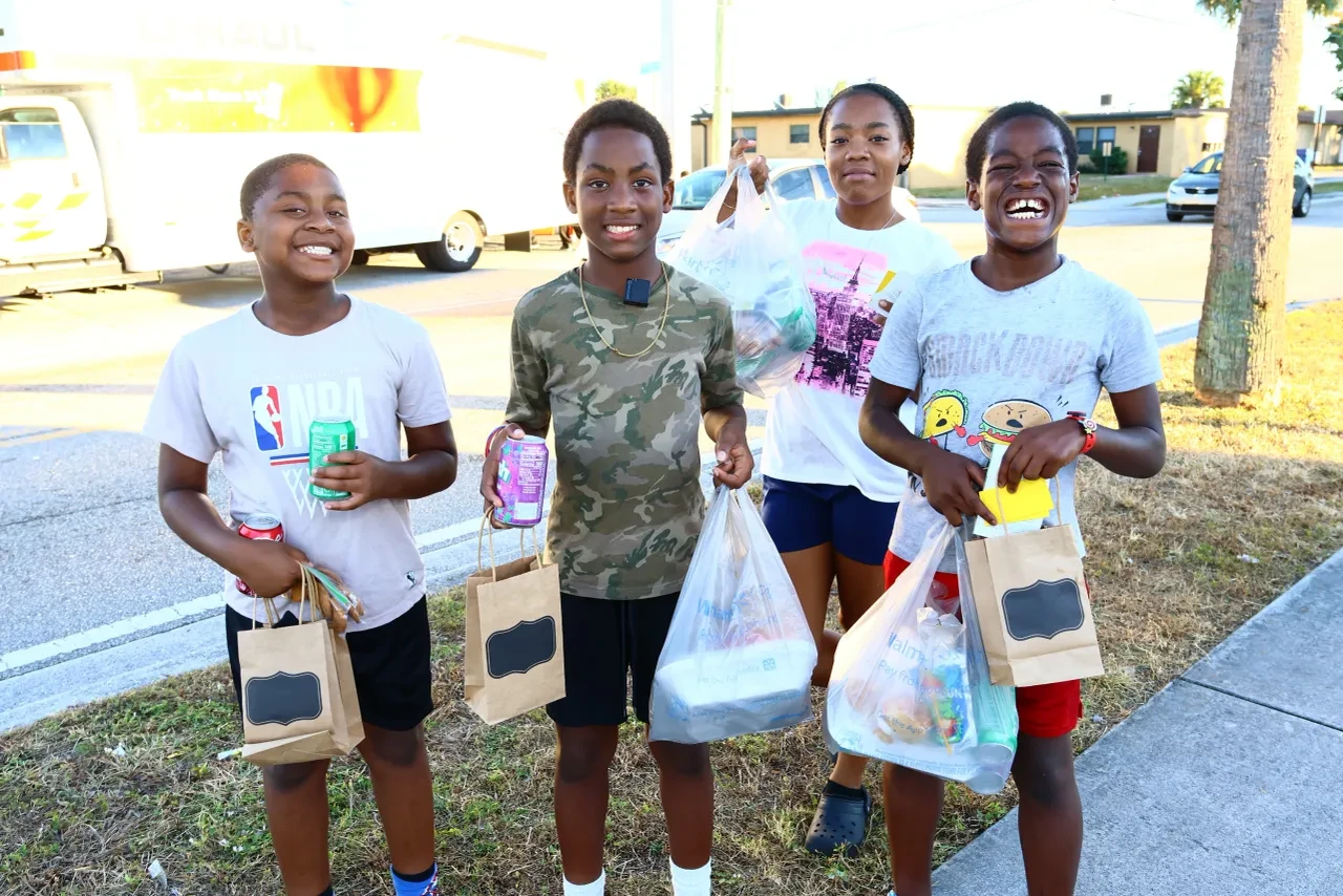 Four smiling children holding donation bags and drinks at outdoor event