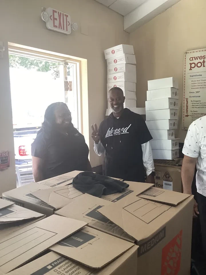 Two volunteers smiling beside stacked Golden Corral food boxes indoors